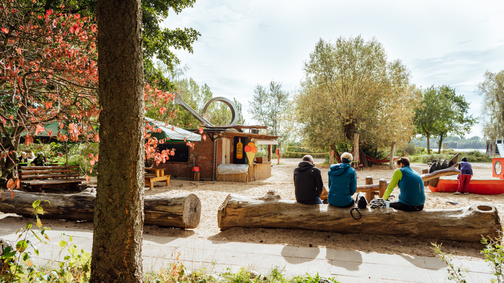 Drei Personen sitzen auf einem Baumstamm im sonnigen Park mit Kiosk und Spielplatz im Hintergrund.