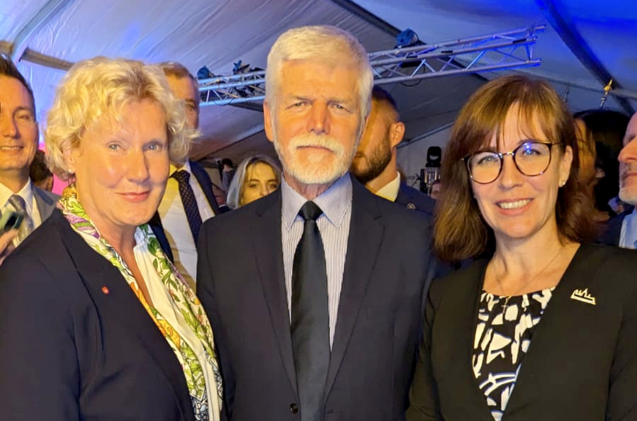 Drei Personen in formeller Kleidung lächeln auf einer Veranstaltung mit mehreren Gästen im Hintergrund.