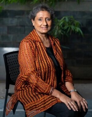 Portrait of a smiling woman with short grey hair sitting on a chair in a patterned orange top.