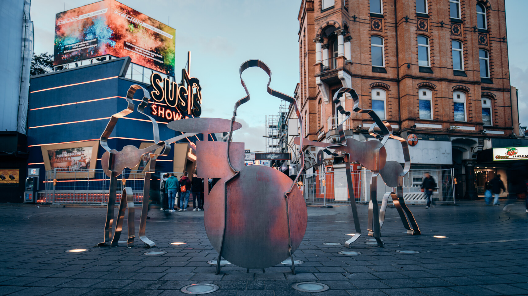 Metallskulptur einer dreiköpfigen Band mit Instrumenten auf einem Platz vor Gebäuden bei Abenddämmerung.