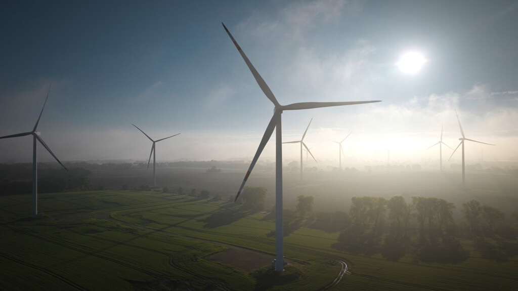 Wind turbines in green fields at a misty sunrise under a blue sky.