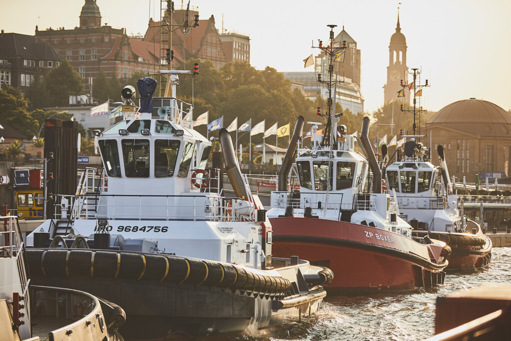 Drei Schlepperboote liegen an einem Hafen mit historischen Gebäuden im Hintergrund bei Sonnenuntergang.