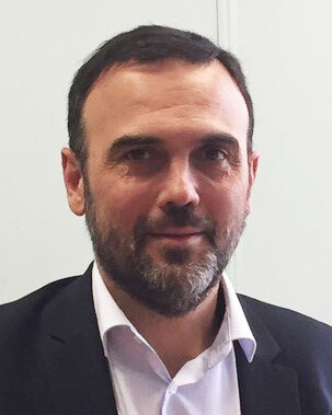 Portrait of a bearded man in a suit jacket and white shirt against a plain background.