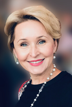 Portrait of a smiling woman with short blonde hair and pearl earrings against a blurred background.