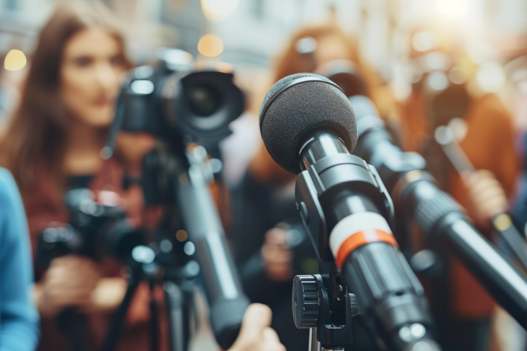 Nahaufnahme von Mikrofonen vor einer verschwommenen Menschenmenge mit Kameras bei einer Pressekonferenz.