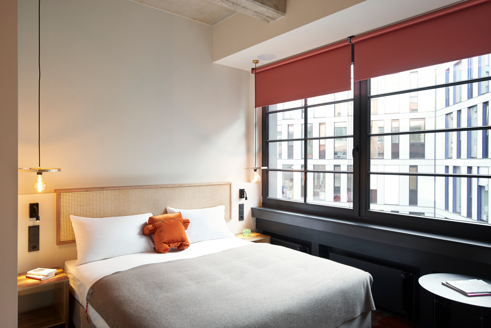 Modernes Schlafzimmer mit großem Bett, zwei Hängelampen und großen Fenstern mit roten Rollos.