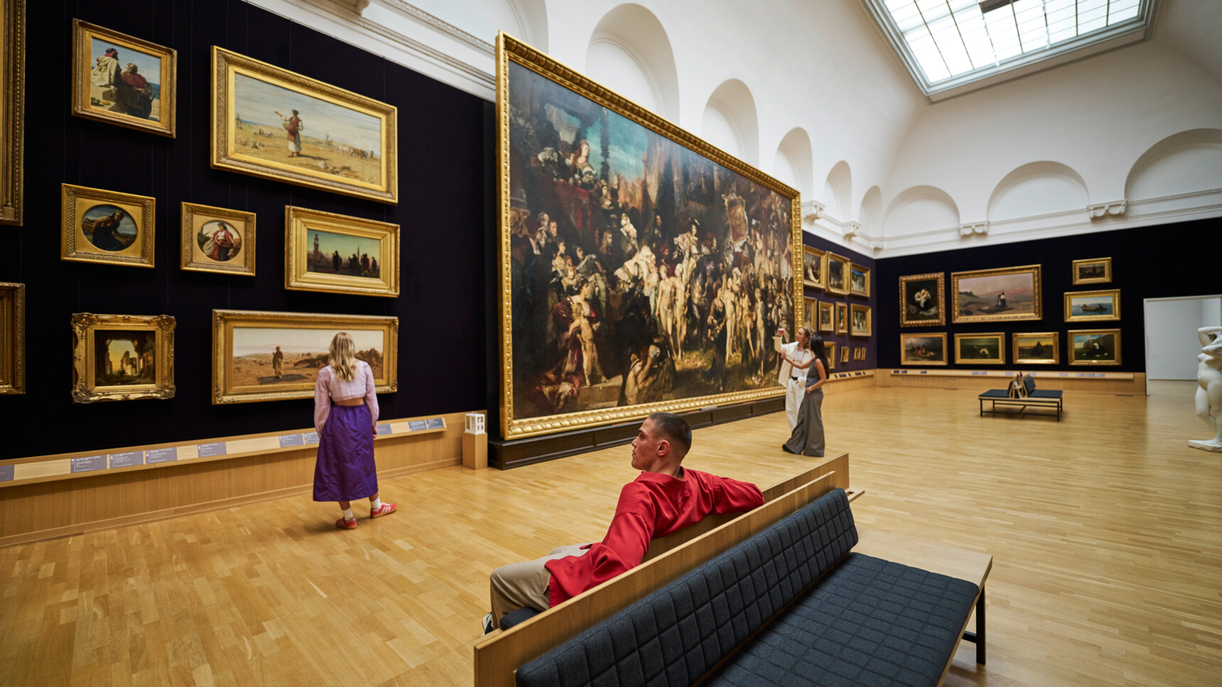 Besucher betrachten Gemälde in einem hellen, geräumigen Kunstmuseum mit Holzboden und hohen Decken.