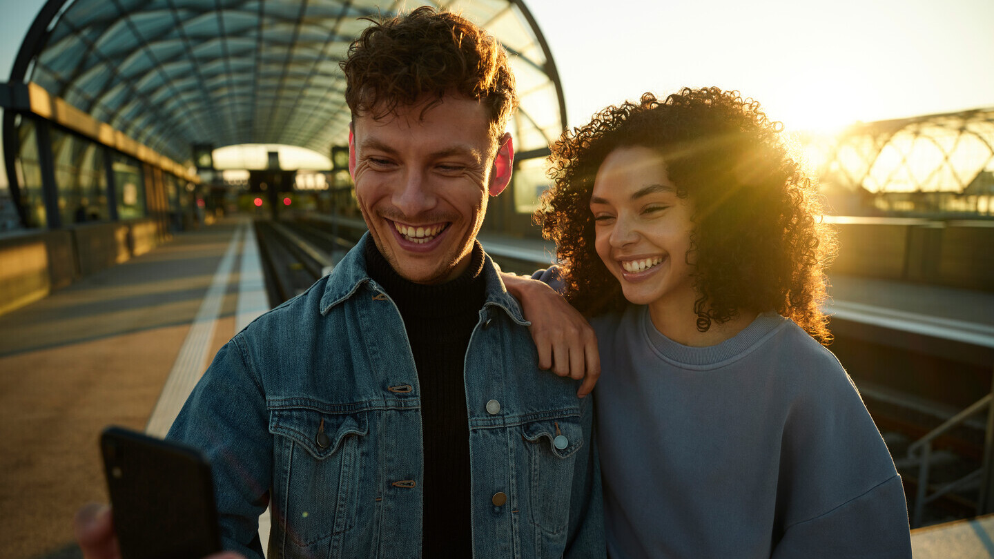 Zwei lachende junge Menschen machen am Bahnhof bei Sonnenuntergang ein Selfie.