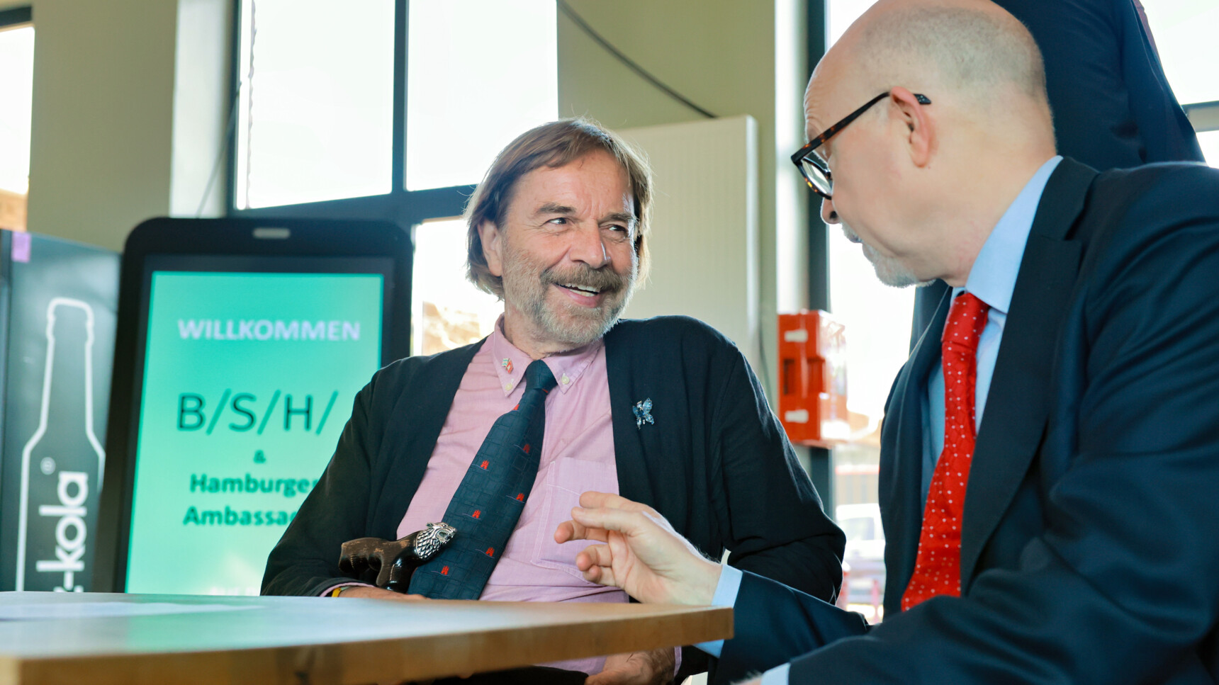 Zwei Männer im Anzug unterhalten sich freundlich an einem Tisch in einem hellen Raum.
