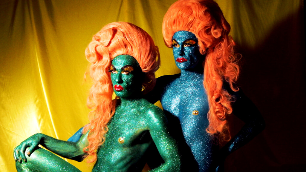 Zwei Personen mit glitzernder blauer und grüner Körperbemalung und orangen, voluminösen Perücken vor gelbem Hintergrund.