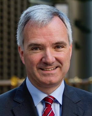 Portrait of a middle-aged man with grey hair wearing a suit and red striped tie, smiling outdoors.