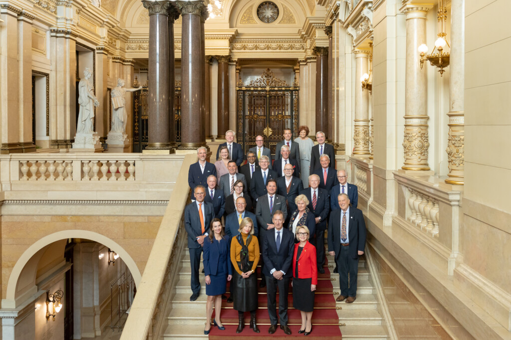 Gruppenfoto von 24 Personen in formeller Kleidung auf einer Treppe in einem prunkvollen historischen Gebäude.