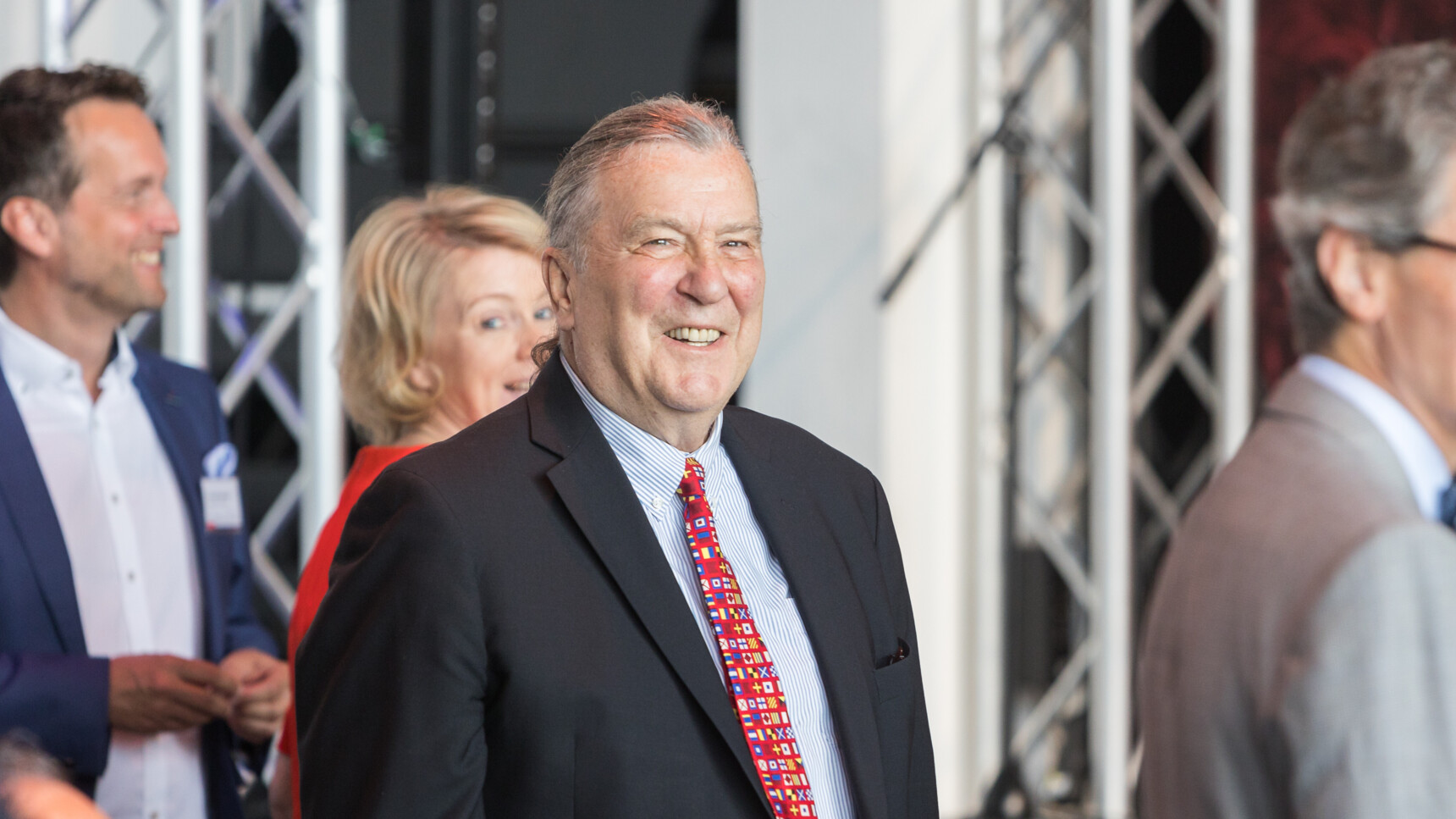 Smiling elderly man in a suit with a colourful tie at a formal event with other attendees in the background.