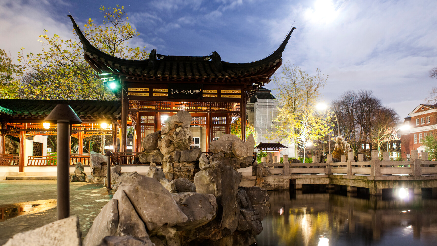 Traditioneller chinesischer Pavillon bei Nacht mit Felsen und Teich im Vordergrund.