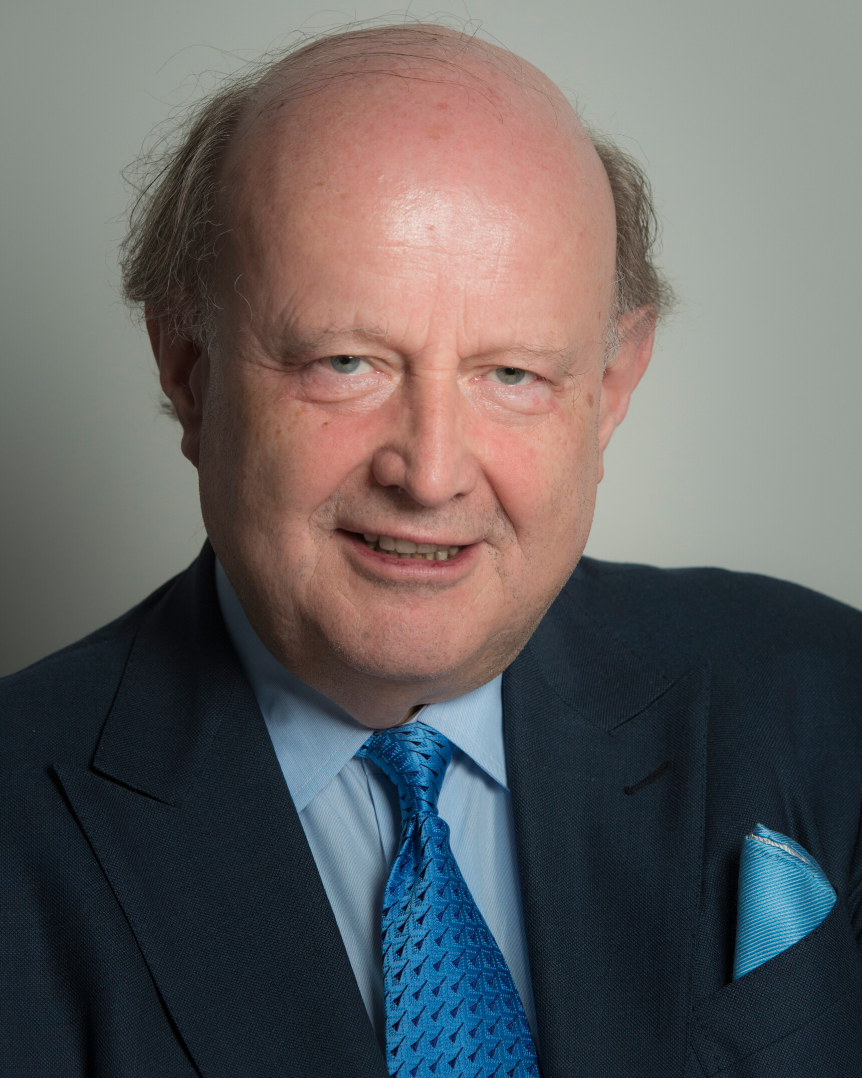 Porträt eines älteren Mannes mit Anzug, blauem Hemd und blauer Krawatte vor grauem Hintergrund.