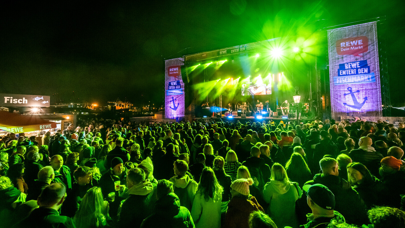 Große Menschenmenge bei einem nächtlichen Open-Air-Konzert mit grüner Bühnenbeleuchtung und REWE-Werbung.