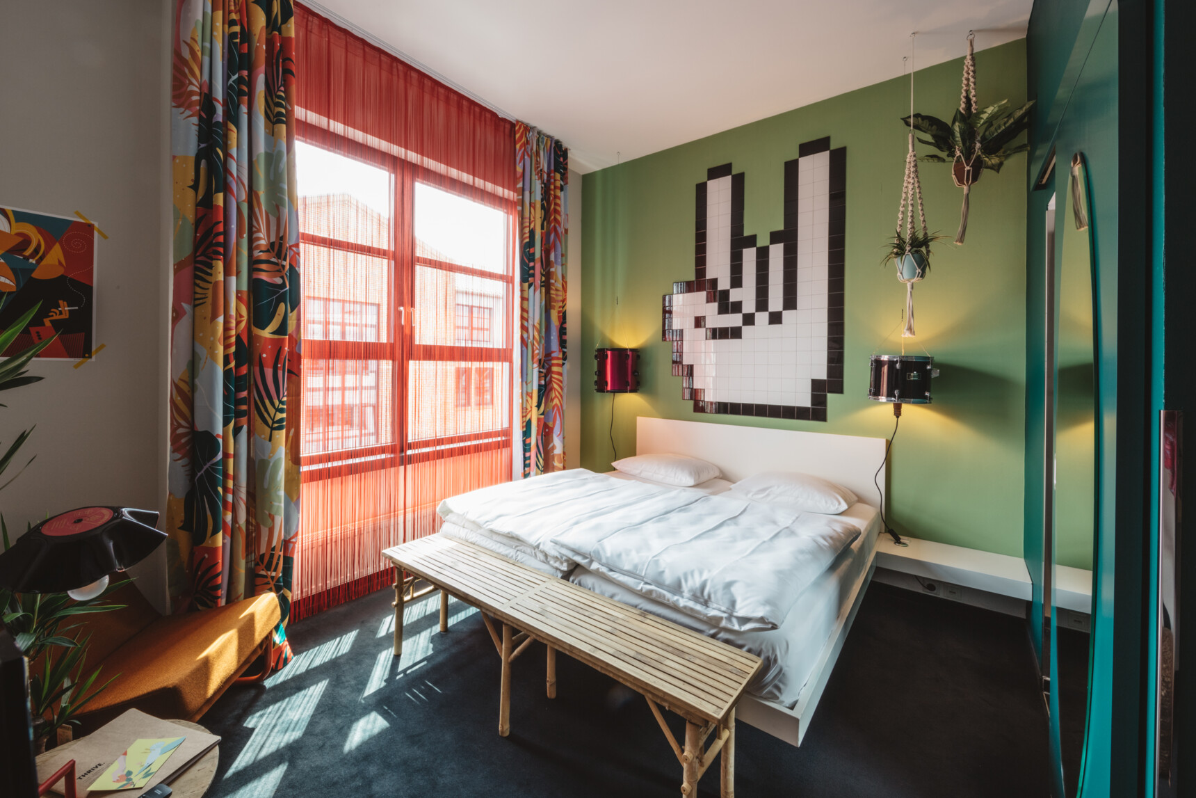 Modernes Schlafzimmer mit grünem Akzentwand, Doppelbett, hängenden Pflanzen und bunter Vorhangdekoration.