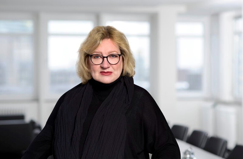 Porträt einer Frau mit blonden Haaren, Brille und schwarzer Kleidung in einem hellen Büro.