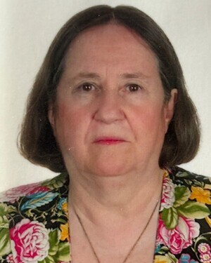 Porträt einer älteren Frau mit braunen Haaren und floral gemustertem Oberteil vor weißem Hintergrund.