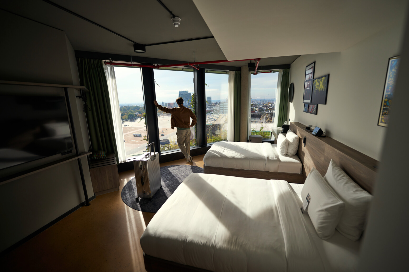 Hotelzimmer mit zwei Betten, einem Koffer und einer Person, die aus großen Fenstern auf die Stadt blickt.