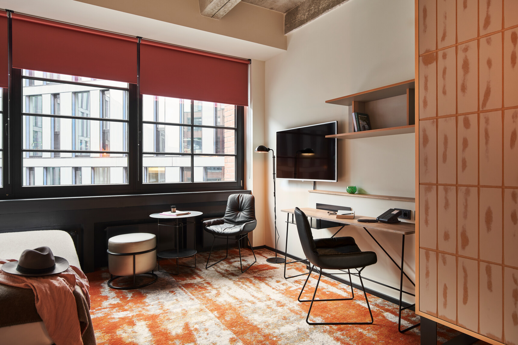 Modern eingerichtetes Hotelzimmer mit Arbeitsbereich, TV, Sitzgelegenheiten und großen Fenstern mit roten Rollos.
