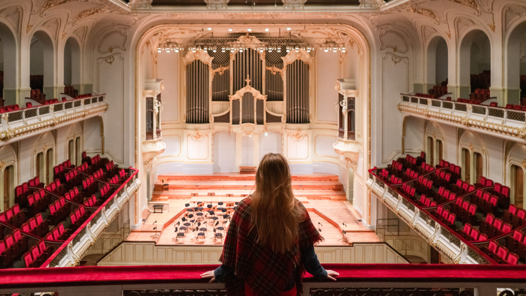 Woman looking from balcony at empty, splendidly furnished concert hall with organ and red seats.