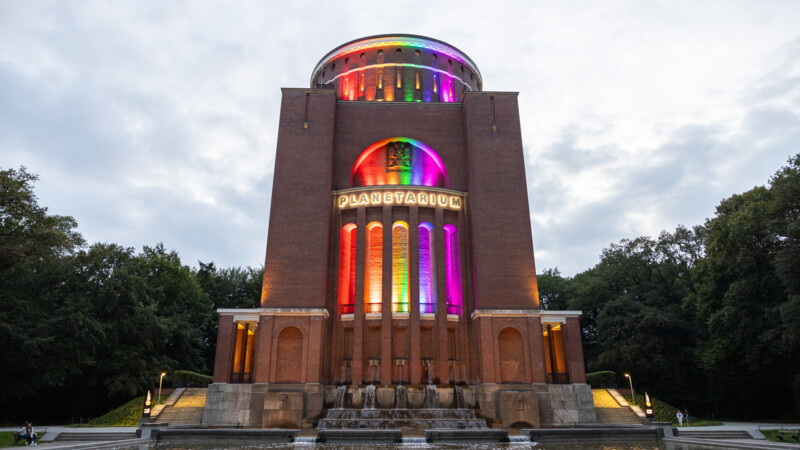 Das Planetarium Hamburg leuchtet abends bunt in Regenbogenfarben mit Wasserfall im Vordergrund.