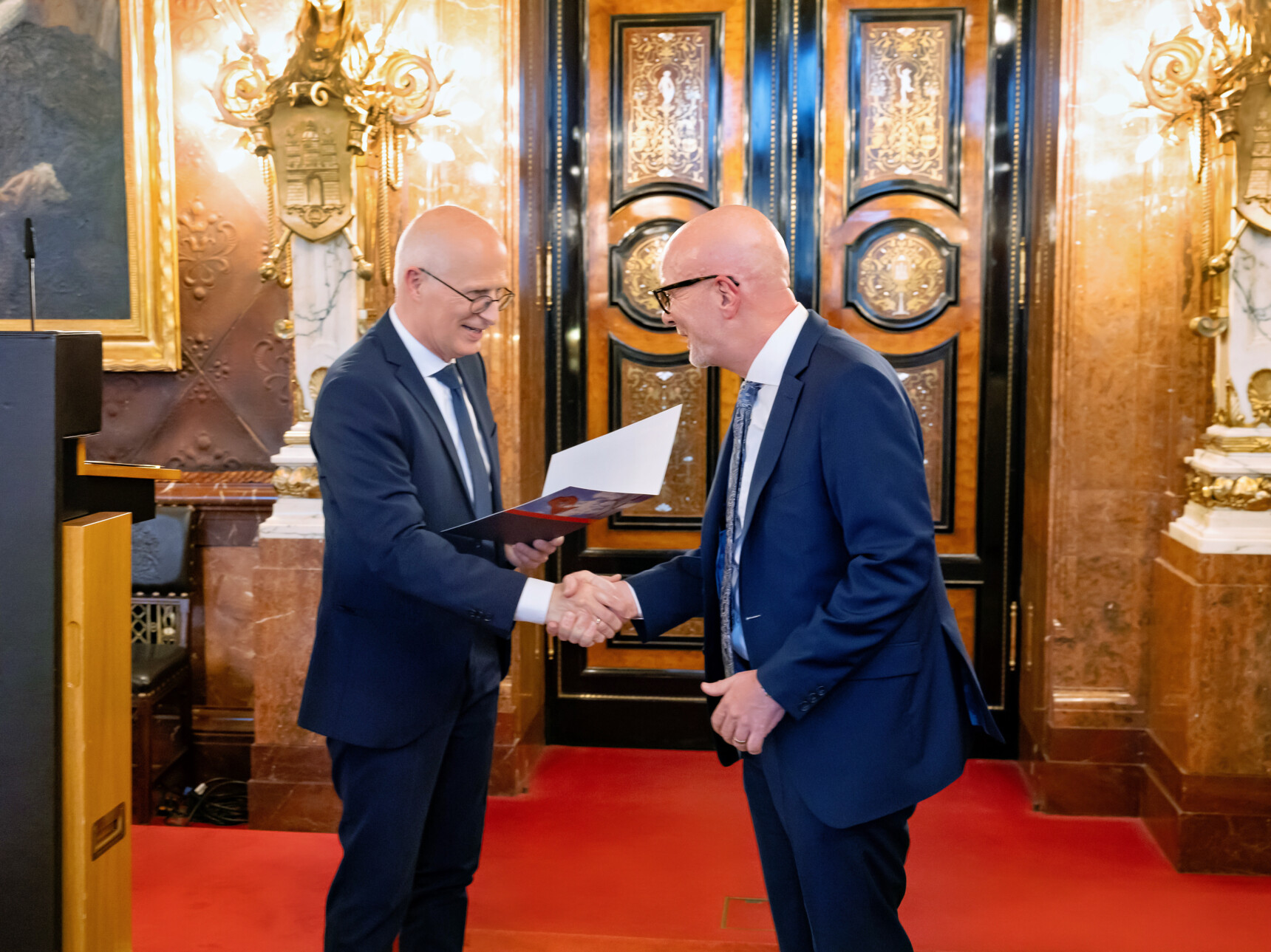 Zwei Männer in Anzügen präsentieren eine Urkunde in einem prunkvollen Raum mit goldverzierten Türen.