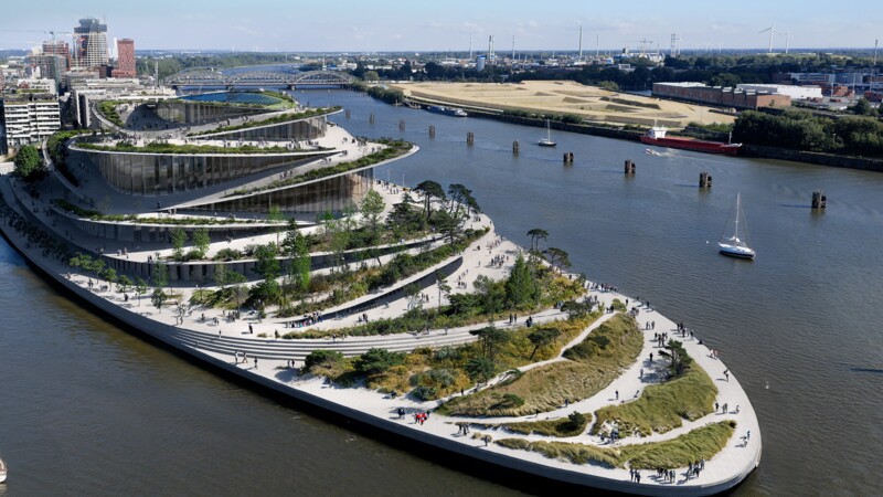 Luftaufnahme einer modernen, terrassenförmigen Parkanlage an einem Fluss mit Spaziergängern und Segelbooten.