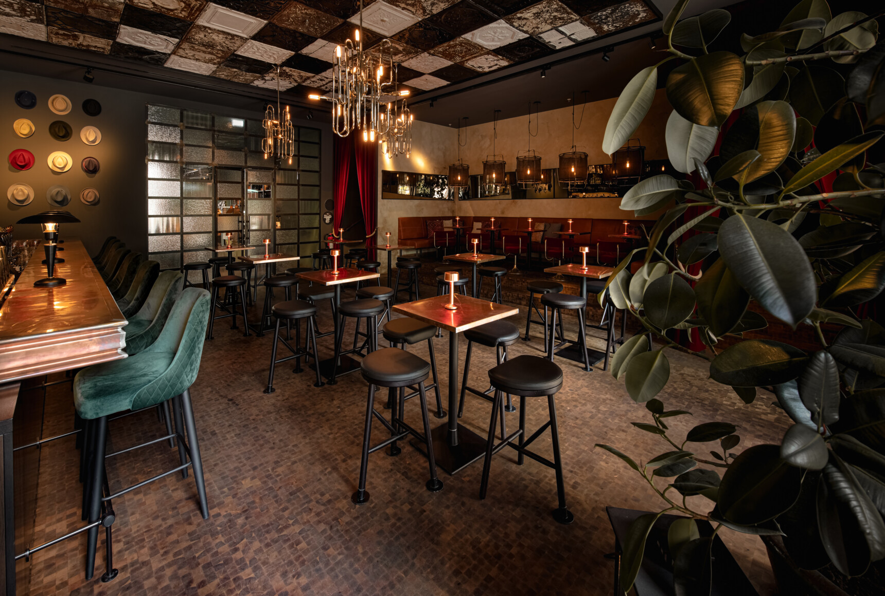 Gemütliche Bar mit hohen Tischen, grünen Barhockern, Kerzenlicht und dekorativen Hüten an der Wand.