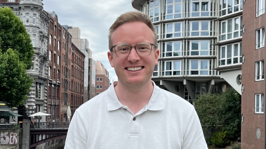 Junger Mann mit Brille und weißem Poloshirt vor urbaner Kulisse mit historischen und modernen Gebäuden.