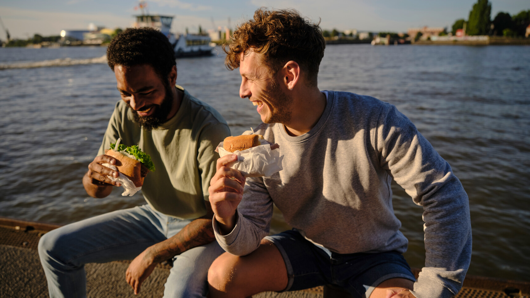 Zwei Männer lachen und essen Sandwiches am Wasser bei sonnigem Wetter.