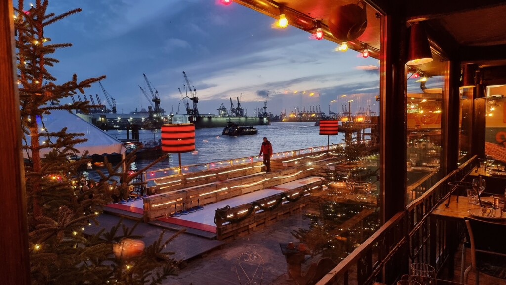 Blick von einem festlich beleuchteten Restaurant auf einen Hafen mit Kränen und einem beleuchteten Steg bei Abenddämmerung.