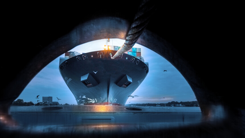 Blick durch eine Öse auf ein angedocktes Containerschiff mit Taube und Seil bei Dämmerung.
