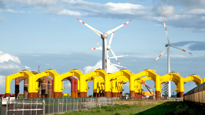 Gelbe Offshore-Windkraftfundamente vor Windrädern unter blauem Himmel auf einer Baustelle.