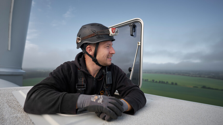 Ein Arbeiter mit Helm und Handschuhen schaut aus dem oberen Zugang einer Windkraftanlage auf die Landschaft.