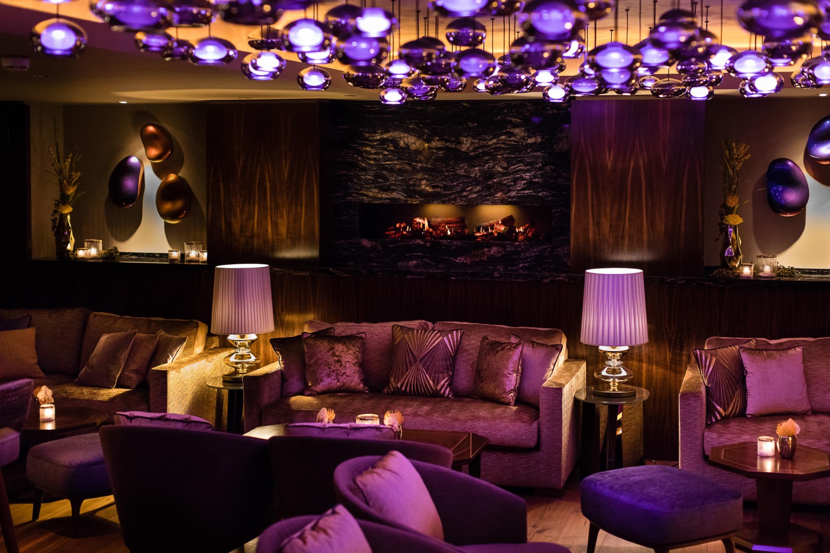 Gemütliche Lounge mit violetten Sofas, warmem Kaminfeuer und moderner Deckenbeleuchtung.