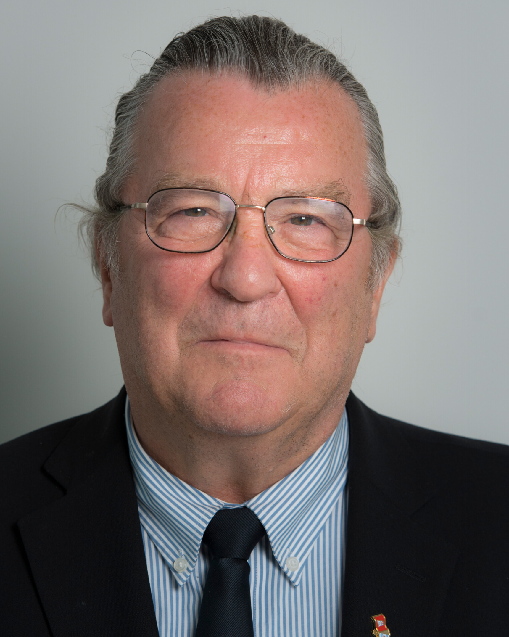 Portrait of an elderly man wearing glasses, a striped shirt, black tie, and dark blazer against a plain background.