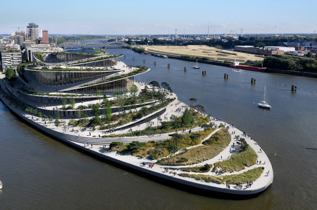 Luftaufnahme einer modernen, terrassenförmigen Parkanlage an einem Flussufer mit Spaziergängern und Segelbooten.