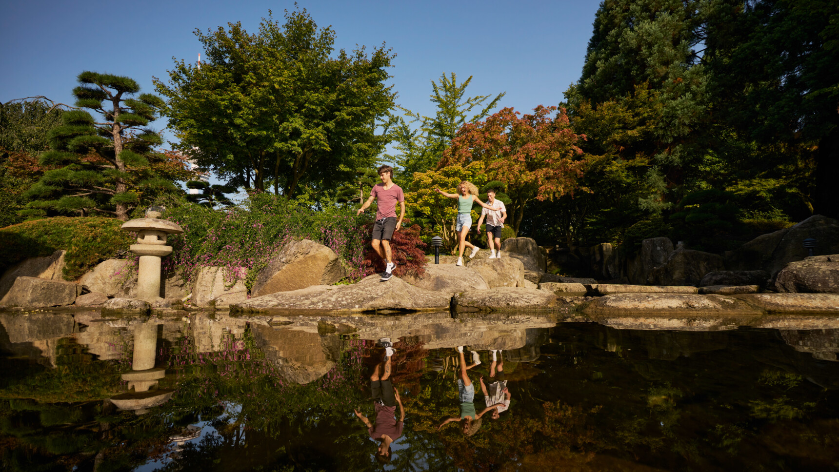Drei junge Menschen springen fröhlich auf Steinen neben einem Teich in einem sonnigen, grünen Garten.