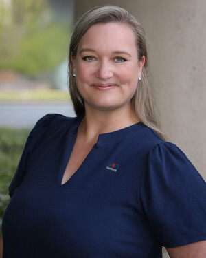 Portrait of a smiling woman with long blonde hair wearing a navy blue top, standing outdoors by a column.