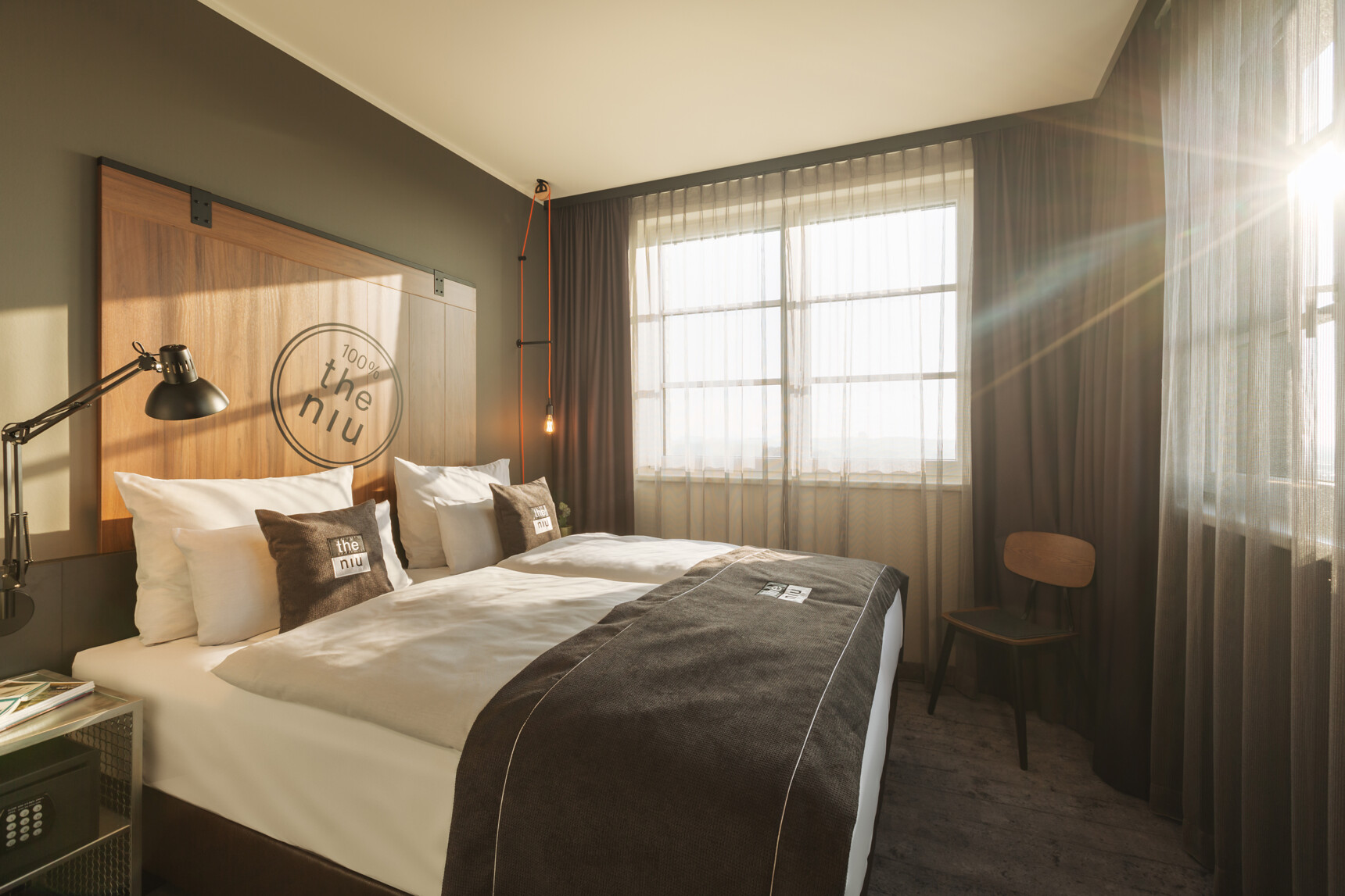 Modern eingerichtetes Hotelzimmer mit Doppelbett, großen Fenstern und Sonnenlicht durch Vorhänge.