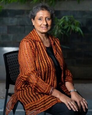 Porträt einer lächelnden Frau mit kurzem grauem Haar, die auf einem Stuhl vor einer grünen Pflanze sitzt.