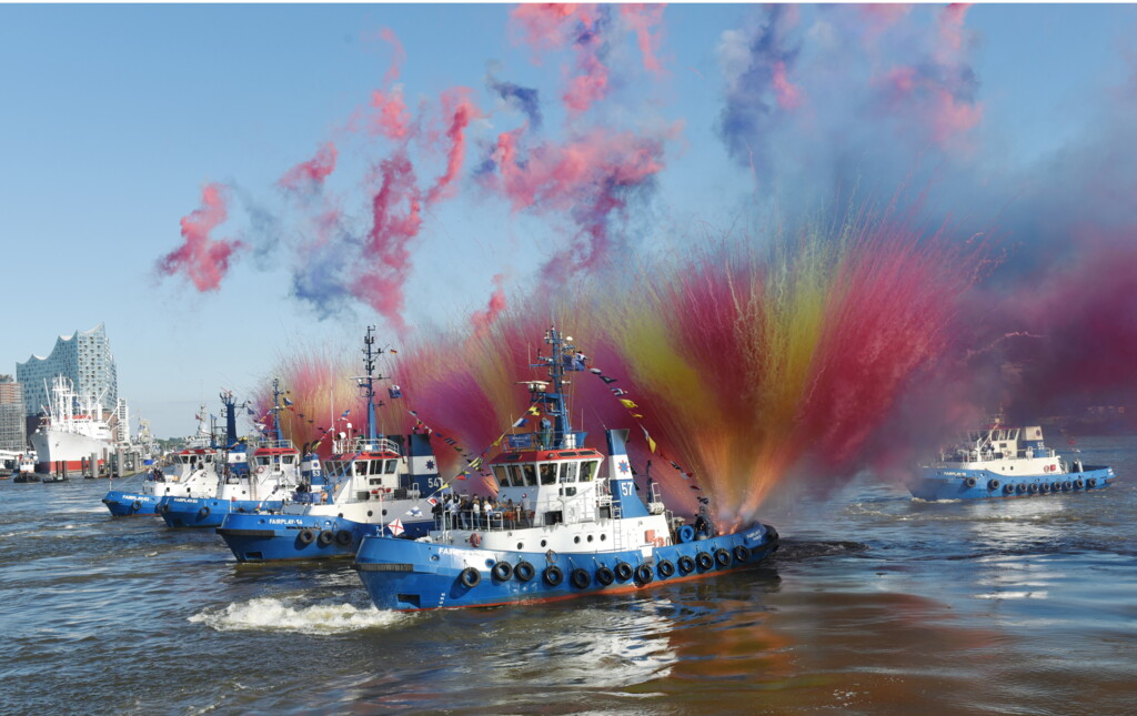 Mehrere Schlepperboote auf dem Wasser, umgeben von buntem Rauch in Blau, Orange und Pink.