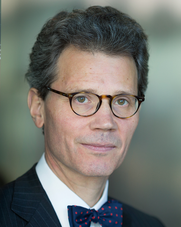 Porträt eines Mannes mit Brille, Anzug und gepunkteter Fliege vor unscharfem Hintergrund.