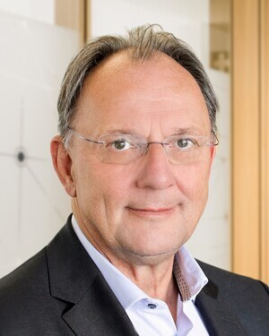 Porträt eines älteren Mannes mit Brille und Anzug vor hellem Hintergrund.