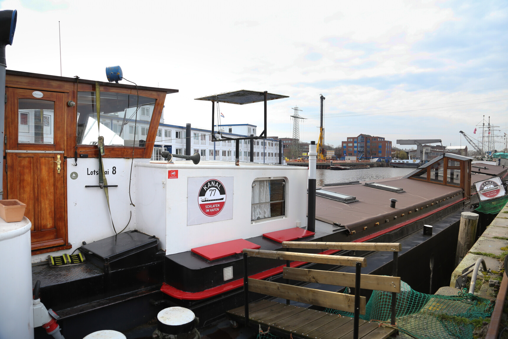 Hausboot mit Holzaufbau und Schild „KANAL 77 Schlafen im Hafen“ am Ufer eines Hafens.