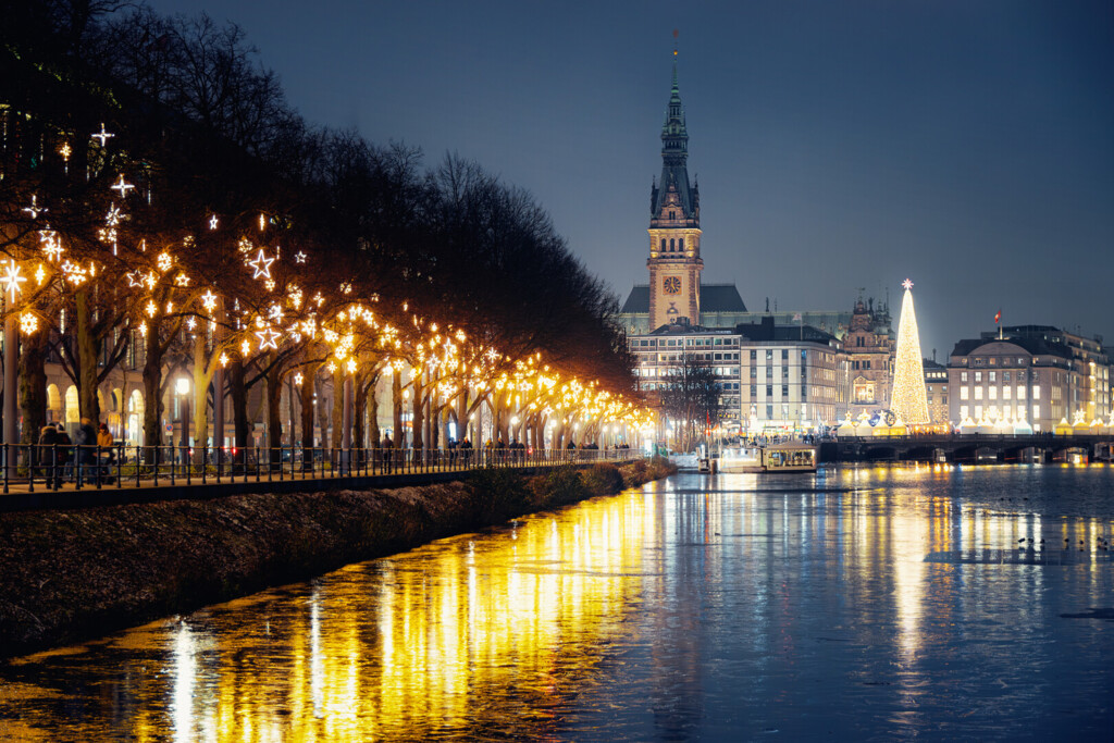 Weihnachtlich beleuchteter Alsteruferweg mit Rathaus und großem Weihnachtsbaum in Hamburg bei Nacht.