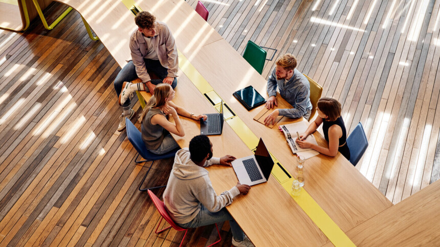 Five young people are sitting at a long wooden table in a modern, light-flooded co-working space. They are engrossed in a lively discussion, working on laptops and taking notes.