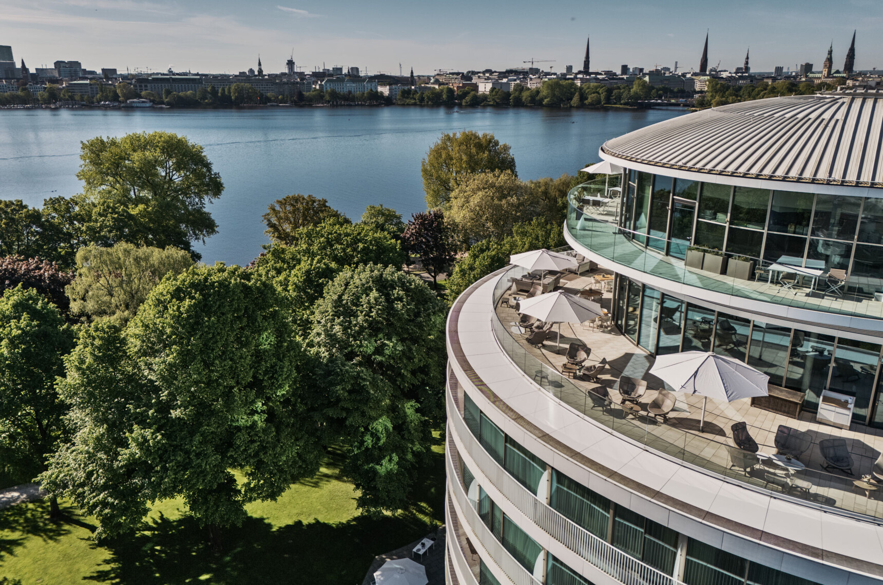 Blick auf Alstersee, grüne Bäume und eine moderne Dachterrasse mit Sonnenschirmen in Hamburg.